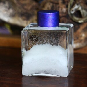 Puede incluir: Una botella de vidrio transparente con polvo blanco en su interior y una tapa azul iridiscente. La botella está sobre una superficie de madera.