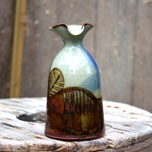 Puede incluir: Un jarrón de cerámica con un esmalte marrón, azul y verde. El jarrón tiene una boca ancha y una base estrecha. Está decorado con un diseño de hojas estilizado.