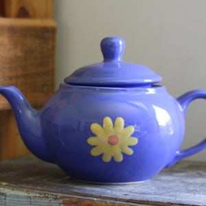 May include: A purple teapot with a yellow daisy design. The teapot has a lid and a handle.