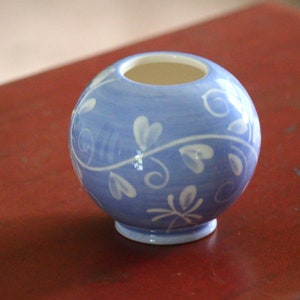 Puede incluir: Un pequeño jarrón redondo de cerámica azul con un diseño floral blanco. El jarrón tiene un acabado liso y brillante y una abertura ligeramente acampanada.