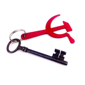 Pode incluir: Um chaveiro de metal vermelho em forma de martelo e foice preso a uma chave esqueleto antiga.