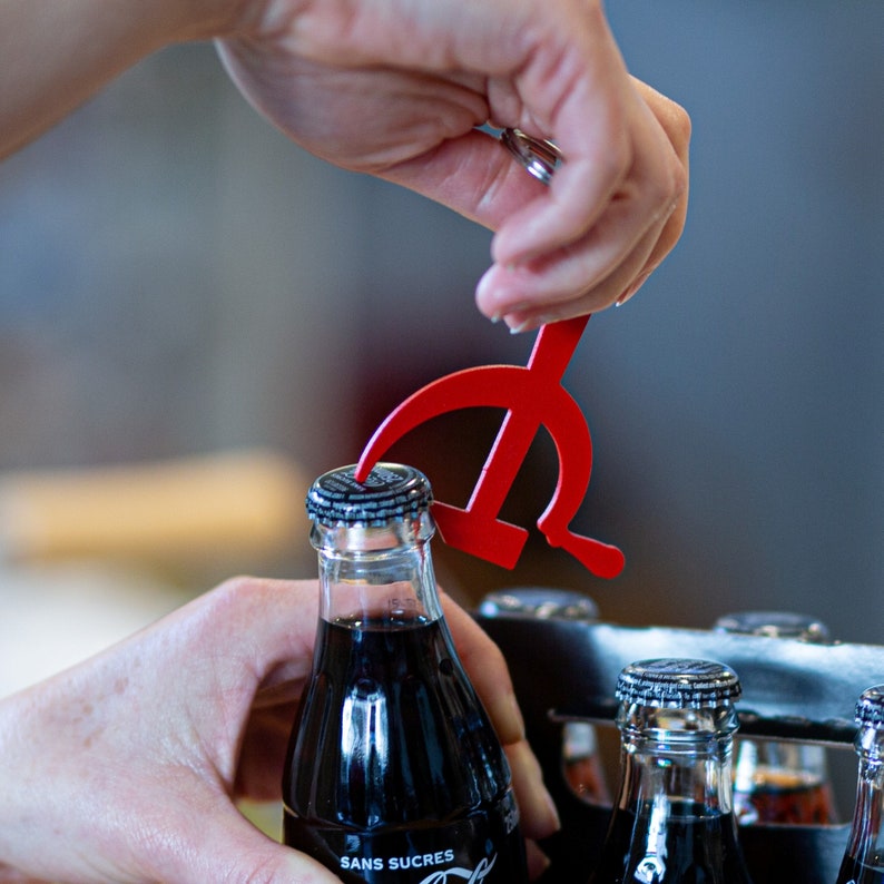 Pode incluir: Um abridor de garrafas de metal vermelho em forma de martelo e foice est&aacute; sendo usado para abrir uma garrafa de cola escura. A garrafa est&aacute; em uma caixa de seis garrafas com outras garrafas.