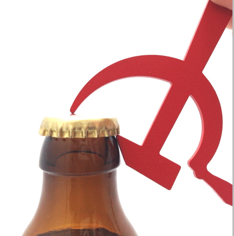 Pode incluir: Um abridor de garrafas de metal vermelho em forma de martelo e foice. O abridor est&aacute; sendo usado para abrir uma garrafa de vidro marrom.