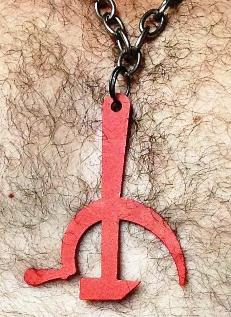 Pode incluir: Um pingente de metal vermelho em forma de martelo e foice, pendurado em uma corrente prateada.