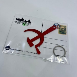 Pode incluir: Chaveiro de metal vermelho em forma de martelo e foice, com um anel de chave prateado. O chaveiro est&aacute; embalado em um cart&atilde;o postal branco com o texto "Design Nicolas Tr&uuml;b X L&eacute;nine - Ref: DCU".