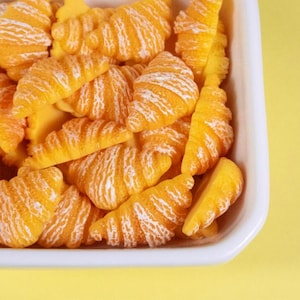 May include: A white rectangular dish filled with miniature, orange-yellow croissants with white, textured detailing. The croissants are arranged in a pile, with some partially visible. The background is a solid yellow.