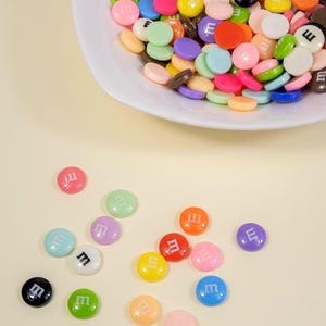 May include: A white bowl filled with colorful, round candies, each with a letter "m" printed on them. The candies are in various colors, including red, blue, green, yellow, and pink. Some candies are scattered around the bowl.