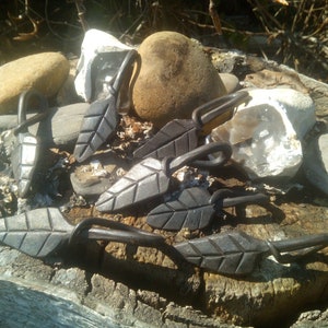 May include: Five black metal leaf-shaped hooks with a twisted stem. The hooks are arranged on a wooden surface with rocks.