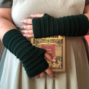 May include: A pair of dark green fingerless gloves with a ribbed knit pattern. The gloves are worn by a person holding a book with the title "César Cabal" on the cover.