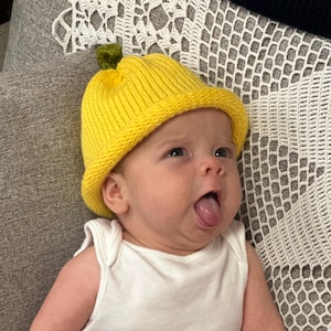 Pode incluir: Um bebé a usar um chapéu de malha amarelo brilhante com um detalhe de caule verde, um body branco com o texto "CLEAN MY PISS?" e um animal de desenho animado, e calças verde claro. O bebé está a mostrar a língua.