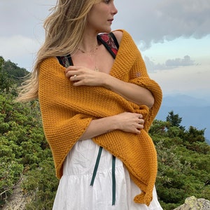 May include: A woman wearing a mustard yellow knitted shawl over a white tiered skirt. She is standing in a mountainous landscape with green trees and a cloudy sky.