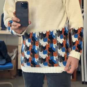 Könnte beinhalten: Ein cremefarbener Pullover mit Rundhalsausschnitt und einem Muster aus bunten Katzen auf der unteren Hälfte. Der Pullover hat lange Ärmel und ist aus weichem, gestricktem Stoff.