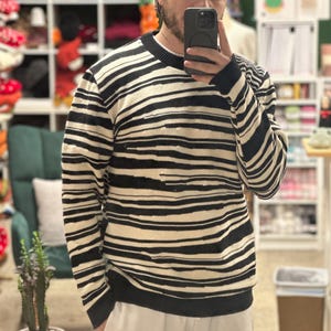 Peut inclure: Un pull à rayures noires et crème. Le pull a un col rond et des manches longues. Les rayures horizontales sont d'épaisseurs variables. Le pull est porté avec un pantalon de couleur claire.