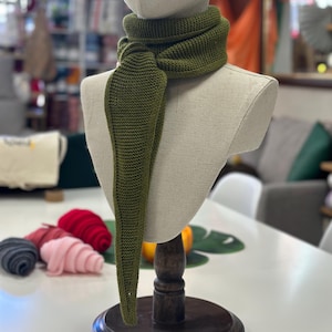 Könnte beinhalten: Ein olivgrüner Strickschal, der auf einer Schaufensterpuppe präsentiert wird. Der Schal ist um den Hals gewickelt und hat ein langes, spitzes Ende. Die Schaufensterpuppe steht auf einem Holzständer, im Hintergrund sind bunte Wollknäuel zu sehen.