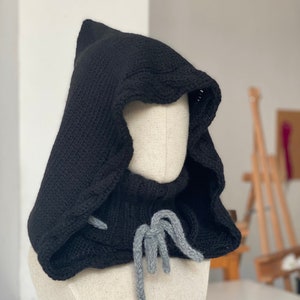 Puede incluir: Bufanda de punto negra con capucha y cordón de ajuste gris. La bufanda está en un maniquí blanco.