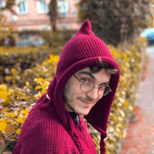 Op de afbeelding: Een persoon draagt een bordeauxrode gebreide trui met capuchon, met een puntige capuchon en een lang, gevlochten koord. De trui heeft een gouden ontwerp aan de voorkant.