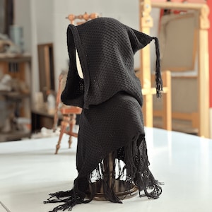 Könnte beinhalten: Schwarzer gestrickter Kapuzen-Schal mit Fransen. Der Schal wird auf einem Holzständer präsentiert.