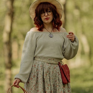 Könnte beinhalten: Eine Frau in einem grauen Pullover, einem Blumenrock und einem Strohhut mit rotem Band geht durch einen Wald und trägt einen Weidenkorb mit roter und weißer karierter Auskleidung.