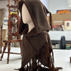 May include: A brown knitted hooded scarf with fringe. The scarf is displayed on a wooden stand.
