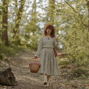 May include: A woman in a forest setting, wearing a straw hat, a grey sweater, a floral skirt, and carrying a wicker basket with a red and white checkered cloth lining.