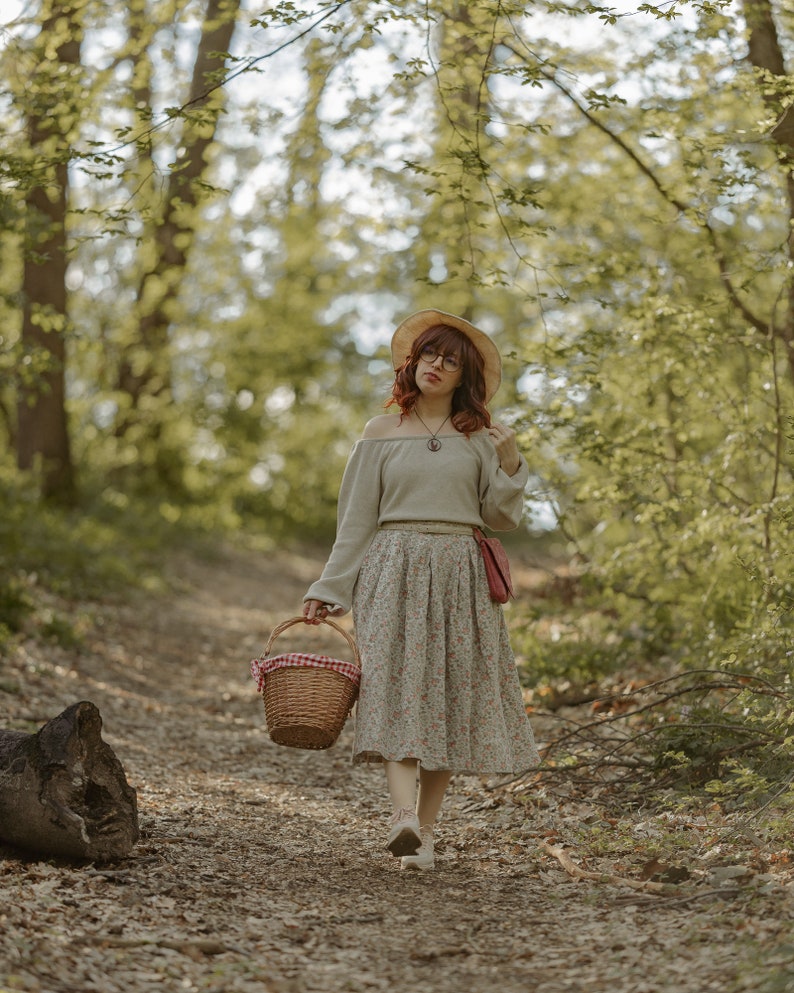 May include: A woman in a forest setting, wearing a straw hat, a grey sweater, a floral skirt, and carrying a wicker basket with a red and white checkered cloth lining.