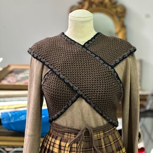 Puede incluir: Un chaleco de punto marrón con ribete negro y gris. El chaleco se lleva sobre una camisa de manga larga marrón y una falda de cuadros marrones y amarillos.