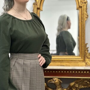 May include: A woman wearing a dark green sweater with a scoop neck and a brown and white plaid skirt. She is standing in front of a mirror.