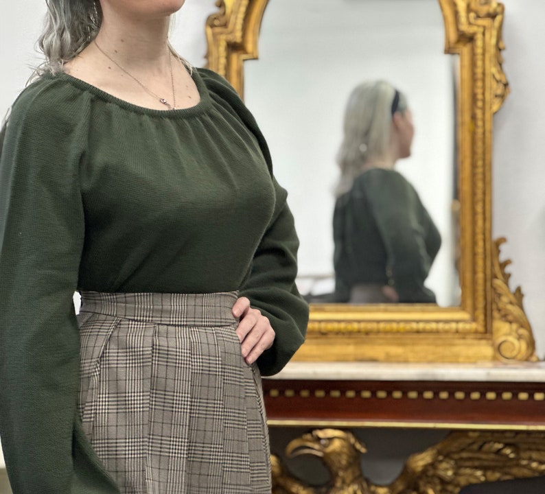 May include: A woman wearing a dark green sweater with a scoop neck and a brown and white plaid skirt. She is standing in front of a mirror.
