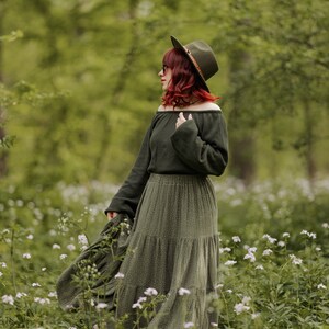 May include: A woman wearing a green off-the-shoulder sweater and a long green tiered skirt with a brown hat stands in a field of white flowers.