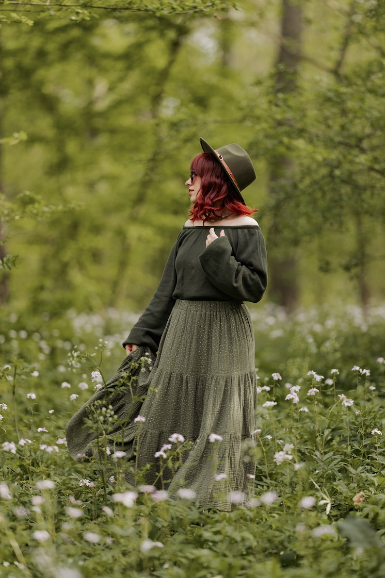 May include: A woman wearing a green off-the-shoulder sweater and a long green tiered skirt with a brown hat stands in a field of white flowers.