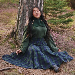 May include: A woman wearing a green sweater and a green plaid skirt sits in a forest setting. She has long black hair and is wearing a green crocheted scarf around her neck.