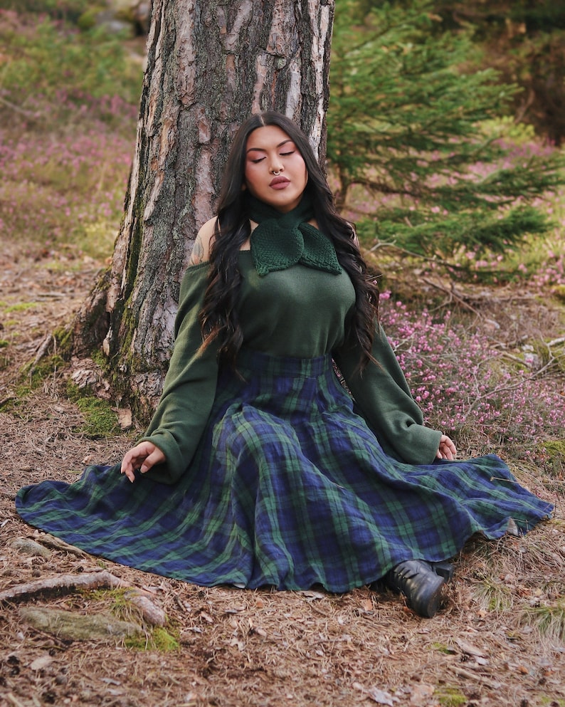 May include: A woman wearing a green sweater and a green plaid skirt sits in a forest setting. She has long black hair and is wearing a green crocheted scarf around her neck.