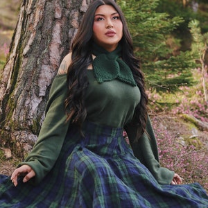 May include: A woman wearing a green sweater with a green bow tie and a plaid skirt sits in front of a tree.