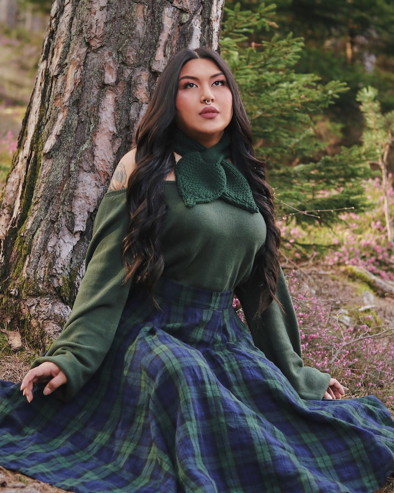 May include: A woman wearing a green sweater with a green bow tie and a plaid skirt sits in front of a tree.
