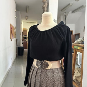May include: A black long-sleeved sweater with a gathered neckline, worn with a white belt and a brown and beige plaid skirt.