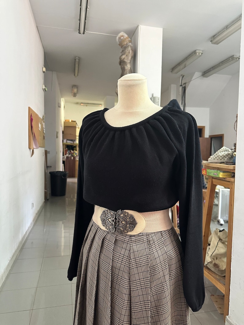 May include: A black long-sleeved sweater with a gathered neckline, worn with a white belt and a brown and beige plaid skirt.