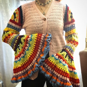 Puede incluir: Un cárdigan de crochet rosa con mangas acampanadas de colores. Las mangas presentan un arcoíris de colores, incluyendo rojo, naranja, amarillo, verde, azul y morado. El cárdigan tiene un cierre de botón.