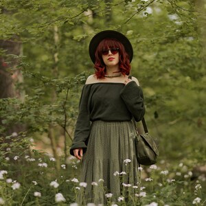 May include: A woman with red hair wearing a green off-the-shoulder sweater, a green skirt with white polka dots, a wide-brimmed hat, and sunglasses. She is standing in a field of white flowers.