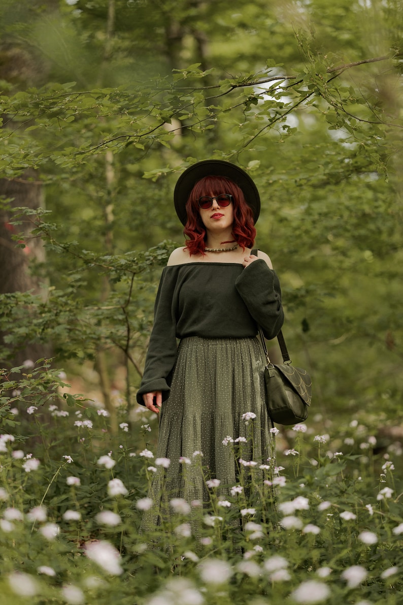 May include: A woman with red hair wearing a green off-the-shoulder sweater, a green skirt with white polka dots, a wide-brimmed hat, and sunglasses. She is standing in a field of white flowers.