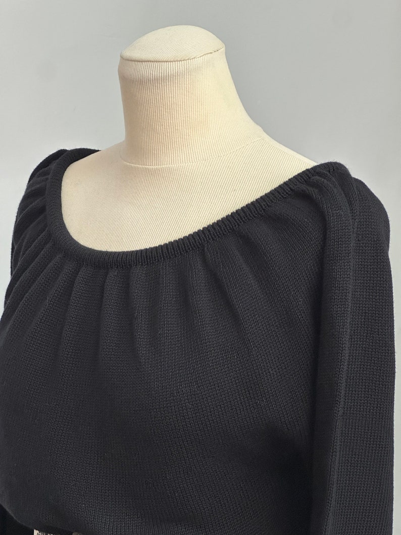 May include: Black knitted sweater with a wide, off-the-shoulder neckline. The sweater has a relaxed fit and a slightly gathered neckline.