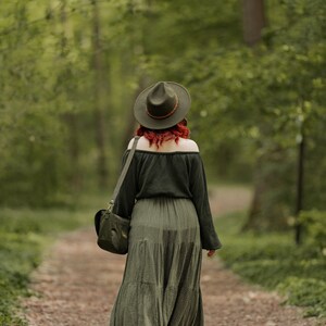 May include: A woman wearing a green dress and a brown hat walks on a path in a forest. She is carrying a brown leather bag over her shoulder.