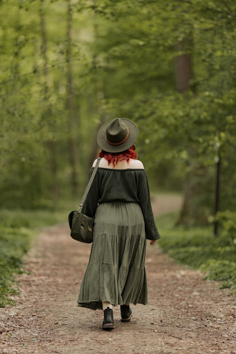 May include: A woman wearing a green dress and a brown hat walks on a path in a forest. She is carrying a brown leather bag over her shoulder.