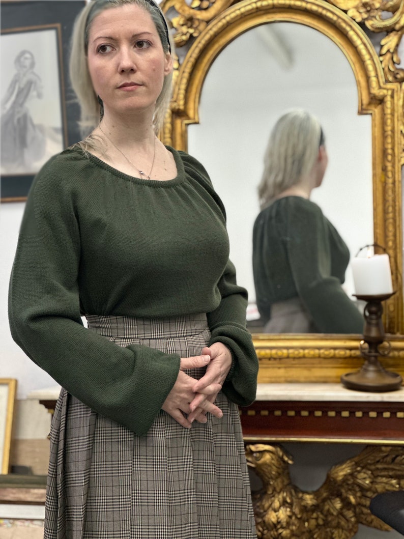 May include: A woman wearing a dark green sweater with a plaid skirt. She is standing in front of a mirror with a gold frame.