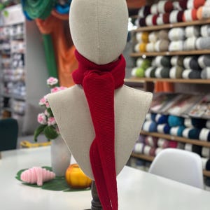 Könnte beinhalten: Ein leuchtend roter Schal aus Rippstrick, der um den Hals einer Schaufensterpuppe drapiert ist. Die Schaufensterpuppe steht auf einem dunklen Holzständer. Im Hintergrund sind Regale mit Garn zu sehen.