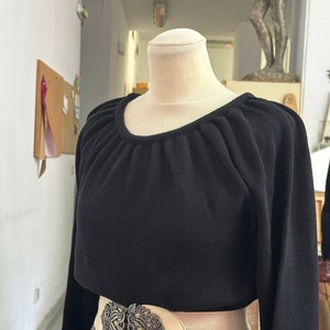 May include: A black long-sleeved sweater with a gathered neckline and a wide, cream-colored belt with a silver ornate buckle. The sweater is worn over a plaid skirt.