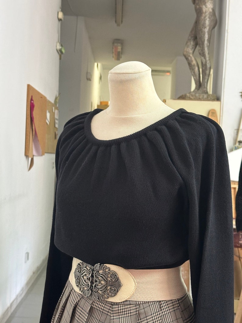 May include: A black long-sleeved sweater with a gathered neckline and a wide, cream-colored belt with a silver ornate buckle. The sweater is worn over a plaid skirt.