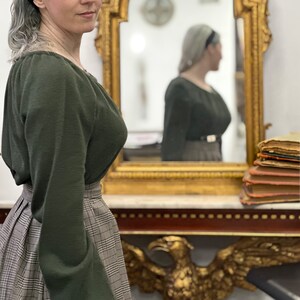 May include: A woman wearing a green long-sleeved sweater with a plaid skirt. She is standing in front of a mirror with a gold frame. The mirror is reflecting a woman wearing a similar outfit.
