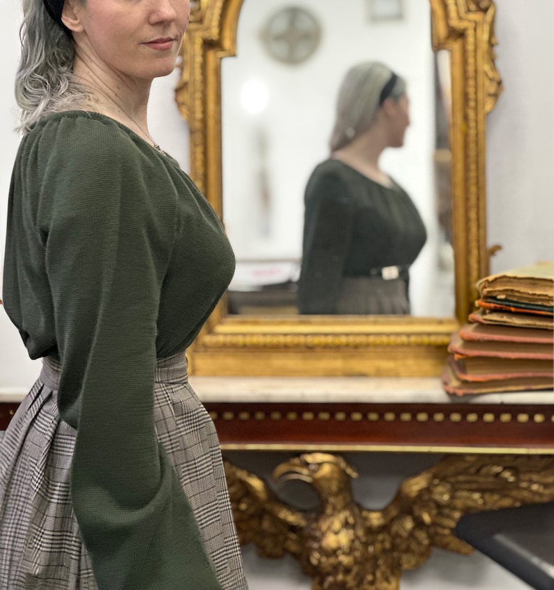 May include: A woman wearing a green long-sleeved sweater with a plaid skirt. She is standing in front of a mirror with a gold frame. The mirror is reflecting a woman wearing a similar outfit.