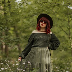 May include: A woman with red hair wearing a green off-the-shoulder sweater, a green tiered skirt, a black hat, and sunglasses stands in a forest setting.