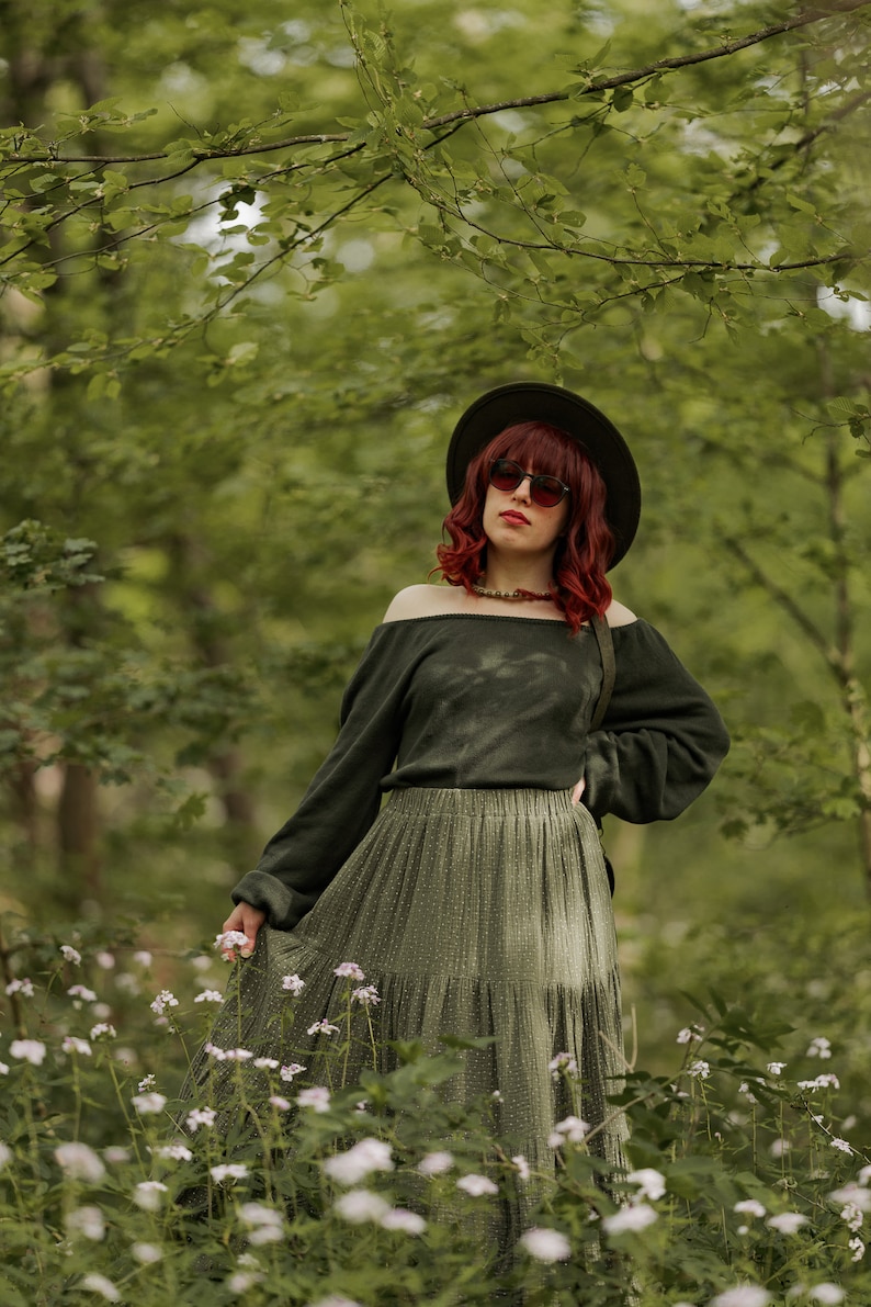 May include: A woman with red hair wearing a green off-the-shoulder sweater, a green tiered skirt, a black hat, and sunglasses stands in a forest setting.